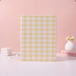 Op de afbeelding: Een notitieboek met een geel en wit ruitpatroon staat op een wit oppervlak. Een kleine witte vaas met bloemen en een roze boek met het woord "Sourdough" staan rechts. Een witte bubbelkaars staat links.