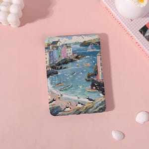 May include: A rectangular case featuring a coastal scene with colorful buildings, boats, and puffins. The artwork depicts a vibrant seaside village with a blue ocean, seagulls, and small boats. The case has a glossy finish and is set against a pink background.