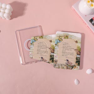 May include: Two rectangular cards with a vintage illustration of a rabbit, cat, and other animals, with text from Beatrix Potter. The cards are in a clear plastic case, with a pink background.