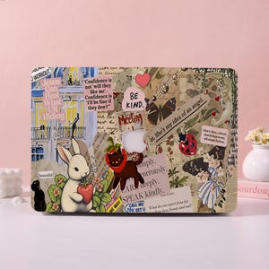 May include: A laptop hard shell case featuring a collage of vintage-style illustrations, including a white rabbit holding a strawberry, a black cat, butterflies, a ladybug, and text snippets like 'BE KIND' and 'Jesus Can Heal What UR Hiding'.