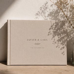 以下が含まれることがあります： ベージュのリネンカバーが付いた横長の写真アルバム。表紙には、JAVIER & LARAというテキスト、小さな葉のイラスト、そして17 DE SEPTIEMBRE 2025という日付が、布地の表面にダークカラーのセリフ体フォントで中央揃えで印刷されています。
