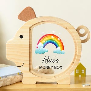 May include: A wooden piggy bank shaped like a pig with a clear front panel. The panel features a rainbow design and the text "Alice's MONEY BOX". The bank is sitting on a wooden shelf with decorative wooden houses and a tree.