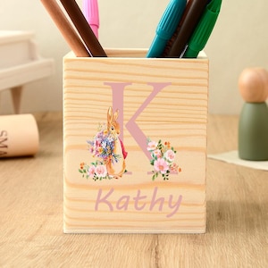 Puede incluir: Un portalápices de madera clara con un diseño de conejito y flores, la letra "K" y el nombre "Kathy" en rosa. El portalápices contiene varios bolígrafos y lápices. El portalápices es de madera clara.