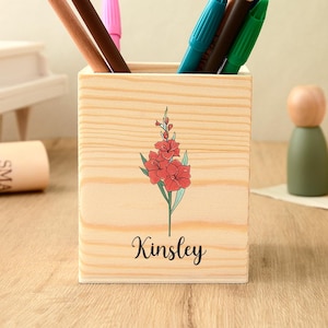 Könnte beinhalten: Ein Holz-Stifthalter mit floralem Design und dem Namen "Kinsley" auf der Vorderseite. Der Halter enthält verschiedene Stifte und Bleistifte. Im Hintergrund befinden sich ein kleines Klavier, eine Pflanze und andere dekorative Gegenstände.