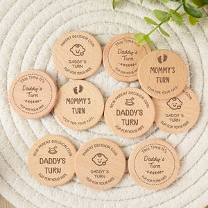 Op de afbeelding: Een verzameling houten beslissingsmunten met gegraveerde tekst en illustraties. De munten bevatten zinnen als "Daddy's Turn" en "Mommy's Turn", samen met baby-thema-iconen. Ze zijn gerangschikt op een witte geweven mat.