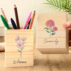 Könnte beinhalten: Zwei Stifthalter aus Holz mit floralen Designs. Einer enthält Stifte und Bleistifte. Der andere wird von einer Hand gehalten. Der erste Halter trägt den Namen "Ariana" und der zweite das Wort "Serenity". Ein Kaktus und Grünzeug sind im Hintergrund.