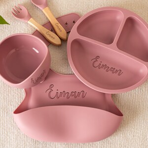 May include: A baby feeding set in dusty rose. The set includes a divided plate, bowl, bib, spoon, and fork. The spoon and fork have wooden handles. The name "Eiman" is engraved on each item. The set is designed for a baby.