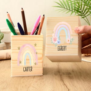 May include: Two wooden pencil holders with rainbow and star designs. One holder is filled with pencils and pens. The other holder has the name "EGBERT" printed on it. The first holder has the name "CARTER" printed on it.