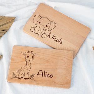Puede incluir: Dos tablas de cortar de madera con diseños grabados. Una presenta un elefante con el nombre "Nicole", y la otra una jirafa con el nombre "Alice". Las tablas son de color marrón claro y tienen un acabado liso, ideales para la cocina o como objetos decorativos.