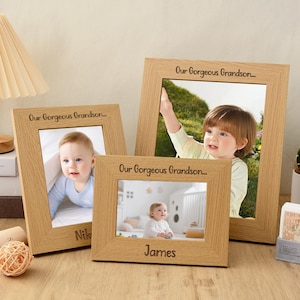 Puede incluir: Tres marcos de fotos de madera clara con fotos de bebé. Los marcos tienen grabada la frase "Our Gorgeous Grandson...". El marco más pequeño tiene grabado el nombre "James", y otro tiene "Nik".