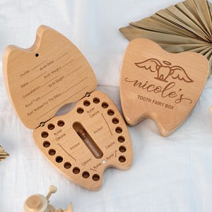 May include: Wooden tooth fairy box with a hinged lid and a compartment for baby teeth. The lid is engraved with the name "nicole's" and "TOOTH FAIRY BOX". The open box has holes for each tooth and a small vial. The open lid has space for baby's information.