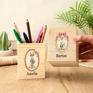 Puede incluir: Dos portalápices de madera con ilustraciones de dibujos animados. Uno presenta un conejo con overoles, el otro un ganso. El primer portalápices está etiquetado como "Amelia", el segundo como "Burton". Varios lápices y bolígrafos de colores están dentro del primero.