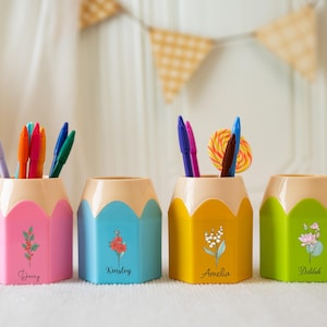 May include: Four pencil-shaped pen holders in pink, blue, yellow, and green, each with a beige top. The holders are decorated with floral designs and names: Daisy, Kinsley, Amelia, and Delilah. Pens and a lollipop are inside.
