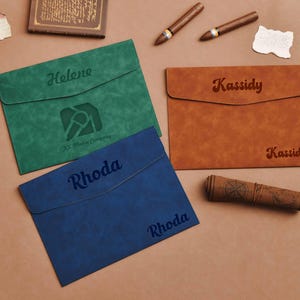 Op de afbeelding: Drie gepersonaliseerde leren documenthouders in groen, blauw en bruin. Elk heeft een naam gegraveerd op de voorkant. Extra items zijn sigaren, een opgerolde kaart en een klein boek.