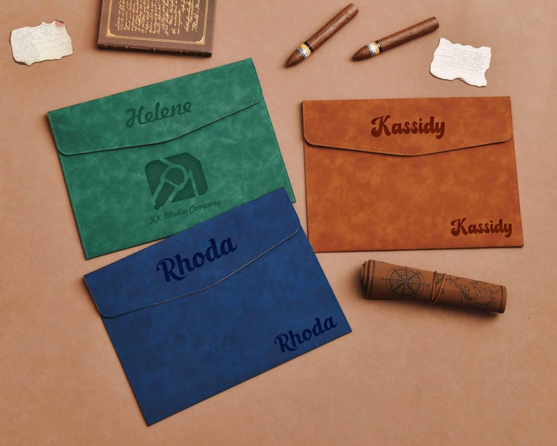 Op de afbeelding: Drie gepersonaliseerde leren documenthouders in groen, blauw en bruin. Elk heeft een naam gegraveerd op de voorkant. Extra items zijn sigaren, een opgerolde kaart en een klein boek.