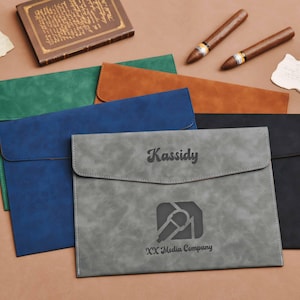 Op de afbeelding: Een verzameling documentenmappen van imitatieleer in verschillende kleuren: groen, blauw, bruin, grijs en zwart. De grijze map is gepersonaliseerd met de naam "Kassidy" en het logo "XX Media Company".