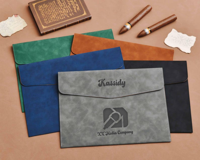Op de afbeelding: Een verzameling documentenmappen van imitatieleer in verschillende kleuren: groen, blauw, bruin, grijs en zwart. De grijze map is gepersonaliseerd met de naam "Kassidy" en het logo "XX Media Company".