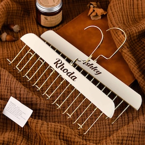 May include: Two off-white wooden tie racks with gold-colored metal bars and hooks. The name "Rhoda" is printed on one rack, and "Ashley" on the other. The racks are on a brown textured surface.
