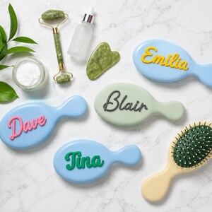 May include: Four oval hairbrushes in light blue, pale green, and cream colors, each featuring a raised name in script font: Emilia in yellow, Blair in dark gray, Dave in pink, and Tina in green. Includes a jade roller, a gua sha tool, and a jar of cream.