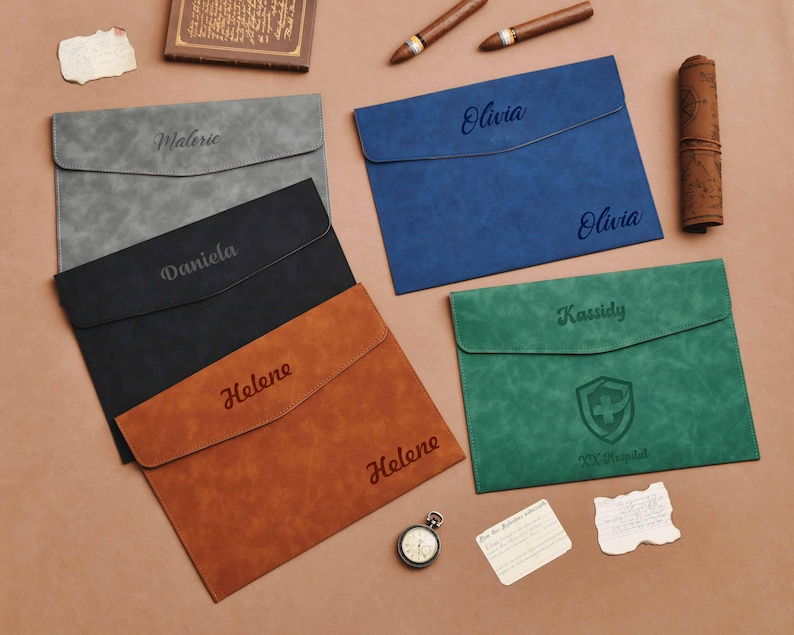 Op de afbeelding: Een verzameling gepersonaliseerde documenthouders in verschillende kleuren: grijs, zwart, bruin, blauw en groen. Elke houder heeft een naam of tekst, &eacute;&eacute;n toont een schildembleem. Accessoires: sigaren, een zakhorloge en een opgerold document.