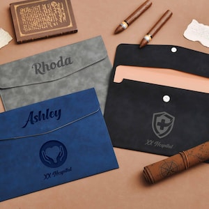 Op de afbeelding: Drie gepersonaliseerde documenthouders in grijs, blauw en zwart. De grijze houder is gegraveerd met "Rhoda", de blauwe met "Ashley" en een hartsymbool, en de zwarte met een medisch embleem. Andere items zijn sigaren en een opgerolde kaart.
