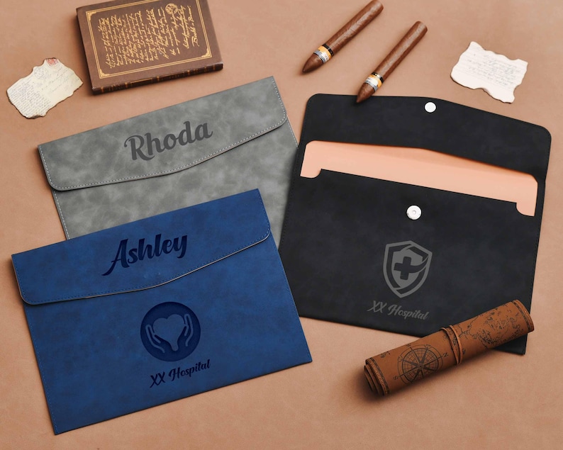 Op de afbeelding: Drie gepersonaliseerde documenthouders in grijs, blauw en zwart. De grijze houder is gegraveerd met "Rhoda", de blauwe met "Ashley" en een hartsymbool, en de zwarte met een medisch embleem. Andere items zijn sigaren en een opgerolde kaart.