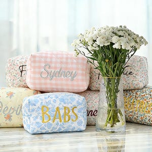 May include: A collection of quilted cosmetic bags in various floral and gingham patterns. Each bag features embroidered names in different fonts and colors. A clear glass vase with white flowers sits in the background, adding a touch of elegance to the display.