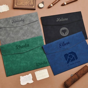 Op de afbeelding: Vier gepersonaliseerde documenthouders in grijs, zwart, groen en blauw. Elk heeft een klepsluiting en een naam of logo gegraveerd op de voorkant. Extra items zijn een horloge, sigaren en een opgerolde kaart.