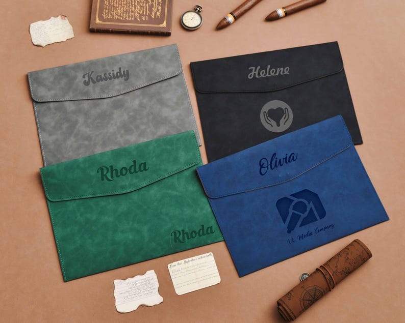 Op de afbeelding: Vier gepersonaliseerde documenthouders in grijs, zwart, groen en blauw. Elk heeft een klepsluiting en een naam of logo gegraveerd op de voorkant. Extra items zijn een horloge, sigaren en een opgerolde kaart.