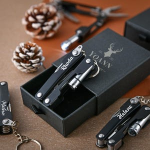 Op de afbeelding: Zwarte multi-tool sleutelhangers met zilveren accenten en gepersonaliseerde namen. Eén sleutelhanger zit in een zwarte geschenkdoos met de naam "YOANN" en een hertenlogo. Andere sleutelhangers worden weergegeven met de namen "Rhoda", "Helene" en "Malerie".