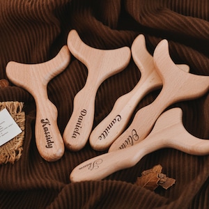 Może przedstawiać: Pięć jasnych, drewnianych narzędzi do masażu z zakrzywionymi uchwytami i unikalnym kształtem. Każde narzędzie ma wygrawerowane imię: Kassidy, Daniela, Camille, Olivia i Sharon. Narzędzia ułożone są na brązowej, teksturowanej powierzchni.