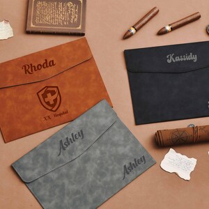 Op de afbeelding: Drie gepersonaliseerde leren documenthouders in bruin, zwart en grijs. De bruine houder heeft een naam en een medisch symbool. De zwarte en grijze houders hebben ook namen. Andere items zijn sigaren, een zakhorloge en een opgerolde kaart.