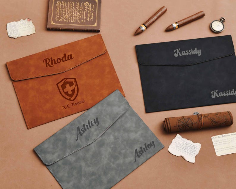 Op de afbeelding: Drie gepersonaliseerde leren documenthouders in bruin, zwart en grijs. De bruine houder heeft een naam en een medisch symbool. De zwarte en grijze houders hebben ook namen. Andere items zijn sigaren, een zakhorloge en een opgerolde kaart.