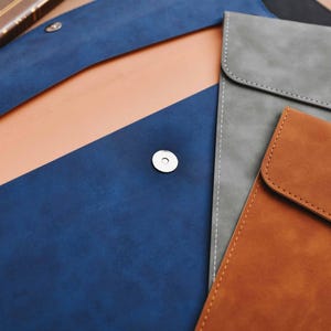 Op de afbeelding: Een assortiment leren laptophulzen in verschillende kleuren. De hoezen zijn in tinten blauw, grijs en bruin, met een magnetische sluiting. De binnenkant van de blauwe hoes is lichtbruin. De hoezen zijn ontworpen met een minimalistische esthetiek.