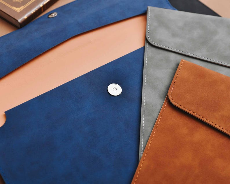 Op de afbeelding: Een assortiment leren laptophulzen in verschillende kleuren. De hoezen zijn in tinten blauw, grijs en bruin, met een magnetische sluiting. De binnenkant van de blauwe hoes is lichtbruin. De hoezen zijn ontworpen met een minimalistische esthetiek.