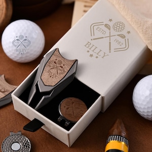 May include: A golf gift set including a divot tool, ball marker, and golf balls. The divot tool and ball marker have a wood grain design with the name "BILLY" engraved. The cream-coloured gift box has the text "ESTD. 1982 BILLY" printed on it.