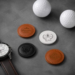 Peut inclure: Quatre marqueurs de balle de golf circulaires en cuir marron, blanc et noir, chacun portant des noms et des motifs gravés au laser. Les marqueurs sont disposés à côté de deux balles de golf blanches alvéolées et d'une montre-bracelet argentée.