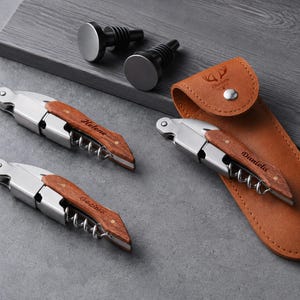 May include: Three stainless steel corkscrews with wooden handles engraved with the names Helene, Gezima, and Daniela, two black metal bottle stoppers, and a tan leather pouch with a snap closure and an engraved deer antler logo.