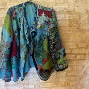 Puede incluir: Chaqueta patchwork en tonos azules, verdes y burdeos colgada de una percha. La chaqueta presenta una variedad de estampados florales y de cachemira. Una aplicación de encaje blanco está cosida en la parte superior del pecho. Mangas largas y cierre de lazo.