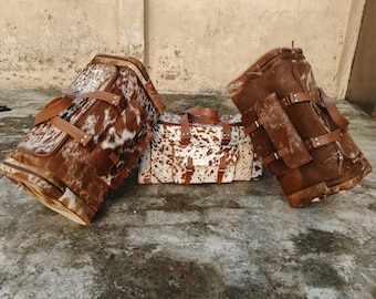 May include: Three brown and white cowhide leather travel bags with brown leather straps and accents. The bags are different sizes and shapes, with a duffel bag in the center and two larger bags on either side.