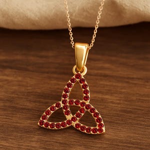 Może przedstawiać: Złoty naszyjnik z wisiorkiem w kształcie węzła Trójcy. Wisiorek zdobiony jest licznymi małymi, okrągłymi, czerwonymi kamieniami szlachetnymi. Naszyjnik ma delikatny łańcuszek i proste zapięcie. Wisiorek ma około 2,5 cm wysokości.