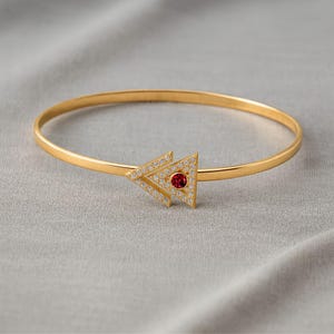 May include: A gold-toned bangle bracelet featuring a central triangular motif. The triangle is outlined with small clear stones and contains a single round, deep red stone at its center. The band is smooth, thin, and circular in shape.