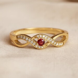 Peut inclure: Une bague en or avec un motif de symbole infini. La bague présente une pierre précieuse rouge ronde centrale, entourée de petites pierres précieuses claires. Le bracelet est de couleur or poli et la bague est posée sur un fond neutre.