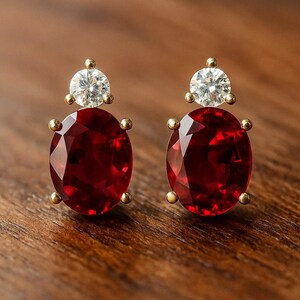 May include: A pair of gold-toned stud earrings. Each earring features a large, oval-cut, deep red gemstone below a smaller, round, clear gemstone. The earrings are set on a wooden surface.