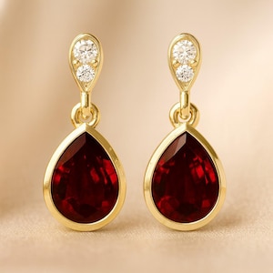 Peut inclure: Une paire de boucles d'oreilles pendantes dorées. Chaque boucle d'oreille présente une pierre précieuse rouge foncé en forme de goutte d'eau, encadrée d'or. Le haut de chaque boucle d'oreille comporte deux petites pierres précieuses rondes et claires.