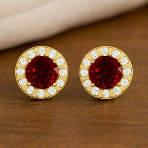 May include: A pair of gold stud earrings with a round, deep red center stone surrounded by a halo of small, white gemstones. The earrings are set on a wooden surface.