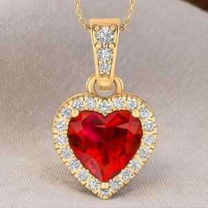Peut inclure: Un collier pendentif doré avec une pierre précieuse rouge en forme de cœur facetté. Le cœur est entouré de petits diamants transparents. La chaîne du collier et la bélière du pendentif sont également dorées.