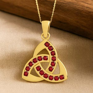 May include: A gold pendant necklace with a Celtic knot design. The pendant is set with multiple red gemstones. The necklace is shown against a neutral fabric, with a wooden backdrop.