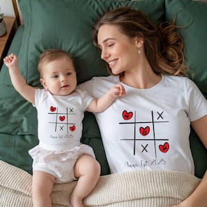 Könnte beinhalten: Passende weiße T-Shirts und ein Body mit einem Tic-Tac-Toe-Design mit roten Herzen und dem Text "Anna Est 2025". Das Baby trägt einen weißen Body mit einer gepunkteten Windel.