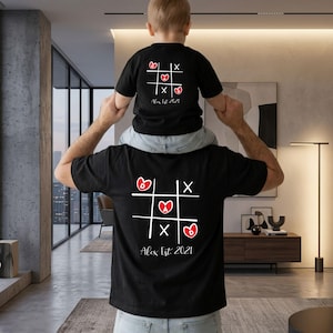 Könnte beinhalten: Schwarze T-Shirts mit einem Tic-Tac-Toe-Design. Das Design enthält Herzen, die Buchstaben "A" und "B" und den Text "Alex Est 2021". Ein T-Shirt wird von einem Kind getragen, das andere von einem Erwachsenen.